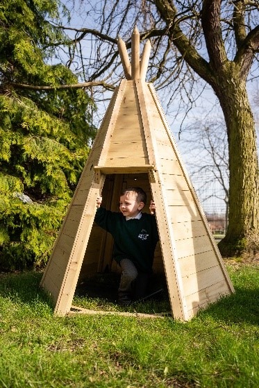 A pupil standing inside a wooden teepee under the trees